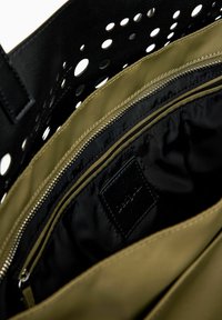 Schwarze, durchlöcherte Lederhandtasche mit olivgrünem Innenfutter, ausgestattet mit einer Reißverschlusstasche und glatter Textur. Das Innere umfasst Details aus schwarzem Stoff.