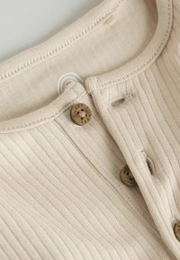 Primer plano de una prenda de punto beige con textura acanalada, botones redondos marrones etiquetados como "NEXT" y ojales visibles.