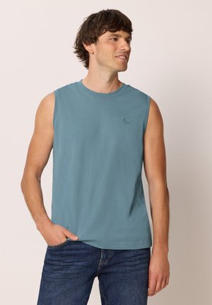 Jeune homme portant un débardeur bleu et un jean, debout avec une main dans la poche, regardant sur le côté, fond clair.