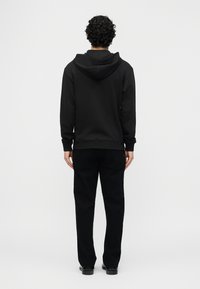 Άνδρας με σγουρά μαλλιά που φοράει μαύρη κουκούλα και μαύρο παντελόνι, στέκεται γυρισμένος με την πλάτη σε απλό ανοιχτό γκρι φόντο.