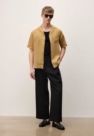 Jonge man met zwarte zonnebril, mosterdkleurig overhemd met korte mouwen over een zwarte top, zwarte broek met wijde pijpen en zwarte loafers, staande op een beige vloer.