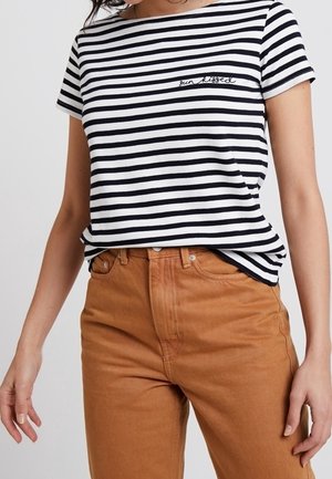 Frau trägt ein schwarz-weiß gestreiftes T-Shirt mit dem Schriftzug „sun kissed“ und hoch taillierte braune Hose, steht mit entspannten Armen.