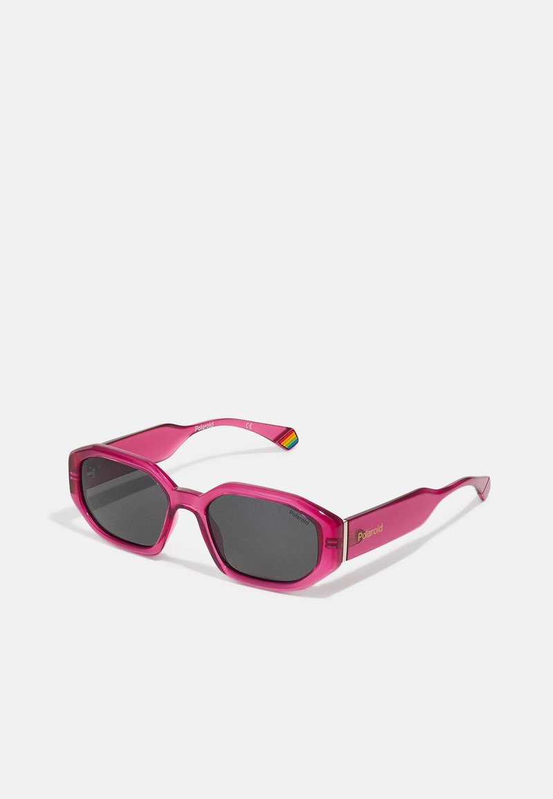 Gafas de sol de plástico rosa con montura angular, lentes oscuros y un logo dorado en el lateral. Una de las varillas presenta un acento arcoíris.