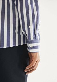 Maglietta a righe viola e bianche in tessuto di cotone, con polsino abbottonato e tasca laterale. Tessuto liscio con silhouette a vestibilità classica.