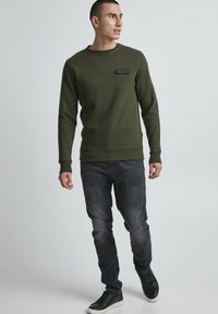 Uomo in piedi che indossa una felpa verde oliva, jeans neri sbiaditi e sneakers nere, che guarda a sinistra contro uno sfondo semplice.