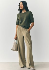 Kvinde iført olivengrøn kortærmet sweater, beige vide bukser, sko med slangeskindsprint, holder en taupe håndtaske, står foran en ensfarvet baggrund.