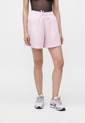 Personne portant un short rose clair à cordon, un haut noir transparent et des baskets blanches Nike, debout devant un fond uni.