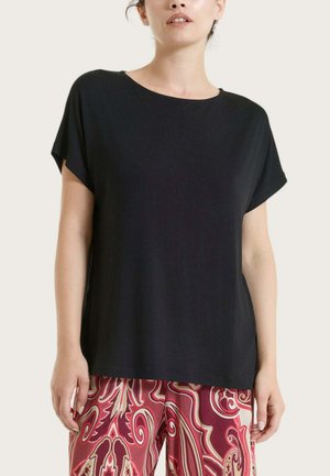 Donna che indossa una maglietta nera semplice a maniche corte e pantaloni rossi con fantasia, in piedi davanti a uno sfondo neutro chiaro.