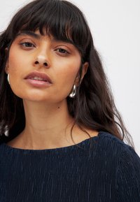 Top texturizado en color azul marino con tela plisada y un escote amplio, acompañado de pendientes redondos de plata brillantes. Piel suave y cabello oscuro.