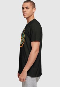 Camiseta de algodón negra con un diseño de escudo multicolor en la parte frontal. Mangas cortas, corte regular, textura suave. Vista lateral mostrada.