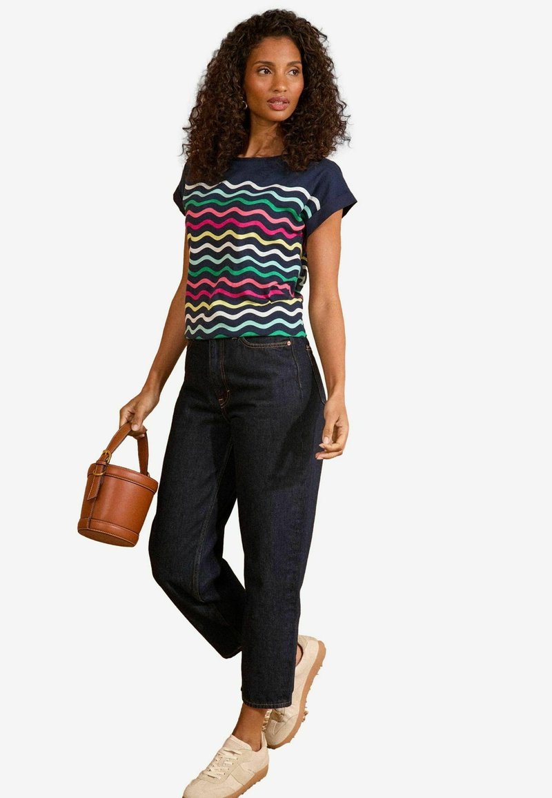 Haut à manches courtes à motifs ondulés multicolores associé à un jean sombre taille haute et des baskets beiges, tenant un petit sac à main marron.