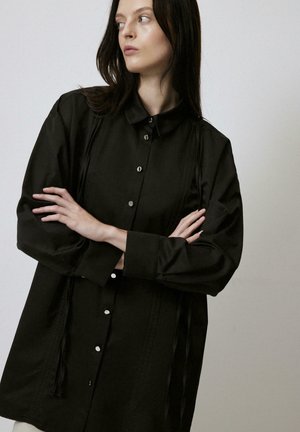 Black button-up shirt with a collar, featuring a relaxed fit, side strings for detail, and smooth fabric texture.