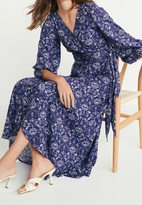 Niebieska kwiatowa sukienka typu wrap z rękawami 3/4, z wiązaniem w talii i zwiewnym dołem. Materiał ma teksturowane wykończenie. Noszona z jasnymi sandałami.