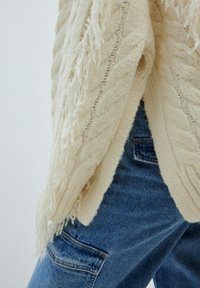 Elfenbeinfarbener, gestrickter Pullover mit strukturiertem Zopfmuster und fransigem Saum, kombiniert mit blauen Jeans aus Denim mit einer Seitentasche.