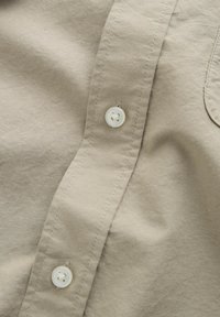 Chemise en tissu beige en gros plan avec deux boutons ronds blancs cousus sur la patte de boutonnage et des détails de couture visibles.