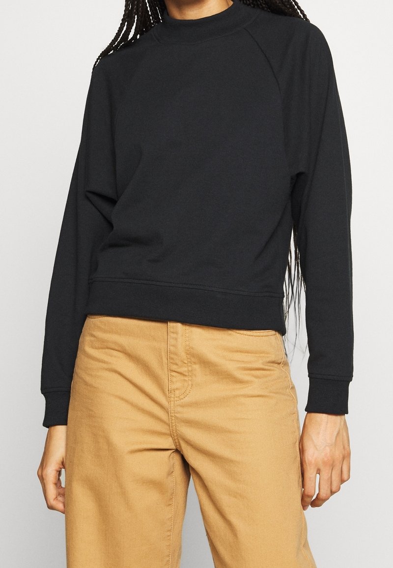Felpa nera con colletto alto e maniche raglan, caratterizzata da una vestibilità comoda e polsini a coste. Abbinata a pantaloni beige.