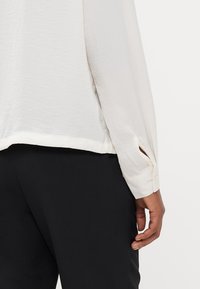 Blouse à manches longues, de couleur crème clair, en tissu lisse, dotée d'une fente sur le côté à l'ourlet et de détails en boutons au niveau des poignets. Portée avec un pantalon noir.