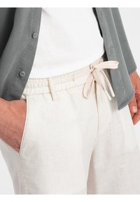 Pantalons en lin beige clair avec une taille élastique et un cordon de serrage, associés à une chemise grise à boutons. Tissu texturé, coupe décontractée.