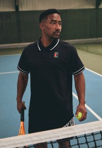 Svart polo skjorte med hvit trim og en liten Nike-logo på brystet, sammen med tennisshorts. Spilleren holder en tenniskølle og en ball.
