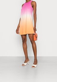 Donna che indossa un vestito ombre da rosa ad arancione senza maniche, mule bianche con tacco e tiene in mano una piccola borsa arancione.