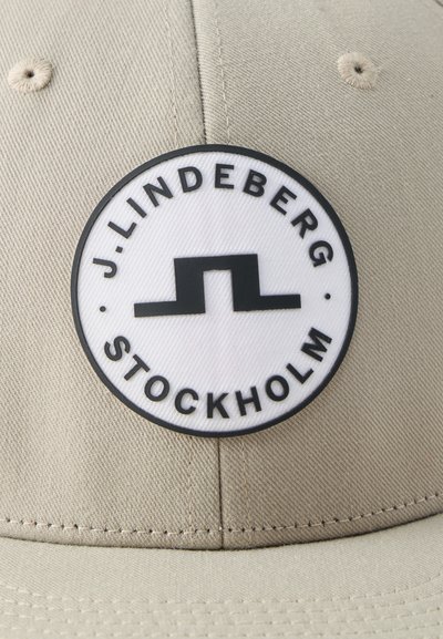 Μπεζ καπέλο με στρογγυλή λευκή ετικέτα που φέρει μαύρο γεωμετρικό λογότυπο και το κείμενο "J.LINDEBERG STOCKHOLM" γύρω από την άκρη.