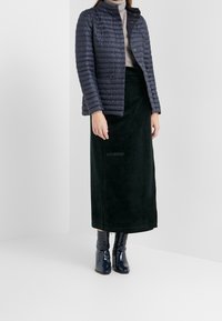 Woman wearing a navy quilted jacket, beige turtleneck sweater, long dark green skirt, and shiny navy ankle boots standing indoors.