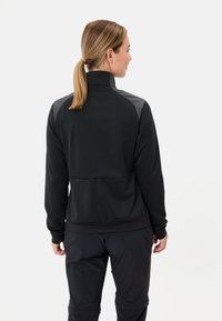 Een vrouw met blond haar in een paardenstaart, gekleed in een zwarte jas met grijze schouderpanelen en zwarte broek, staande tegen een effen witte achtergrond.