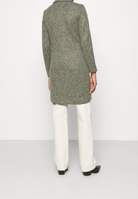 Abrigo largo de punto gris con textura y cuello, que presenta mangas largas y un corte recto, combinado con pantalones blancos y zapatos negros.