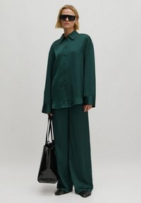 Groene satijnen blouse met een kraag, lange mouwen, gecombineerd met bijpassende wijde broek. Accessoires zijn een zwarte tote bag en overgrote zonnebril.