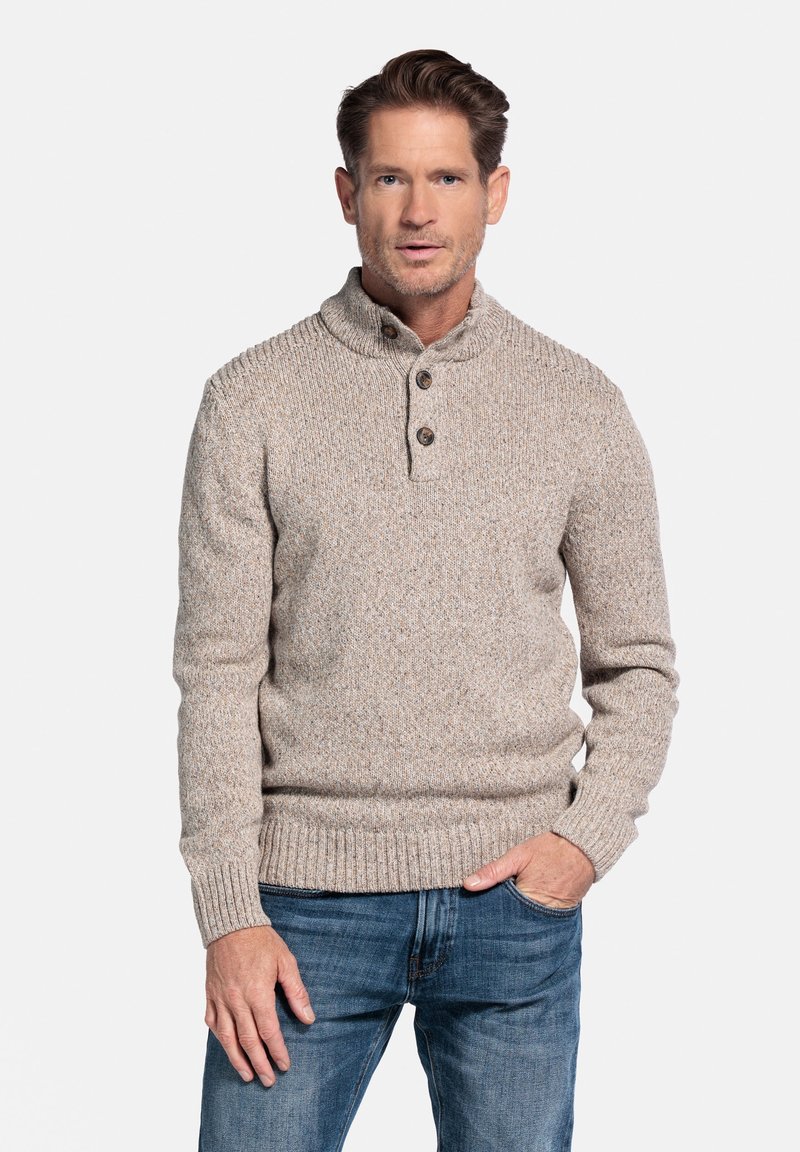 Beige gebreide trui met hoge kraag, twee-knopsluiting en geribbelde manchetten en zoom. De textuur van de stof heeft subtiele spikkels. Draagt met blauwe jeans.