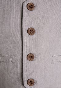 Tissu gris clair avec un tissage texturé. Cinq boutons marron avec un logo gravé. Présente un col et une poche avant sur le côté gauche.