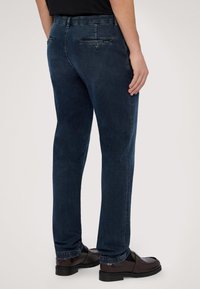 Tmavě modré džínové kalhoty se střihy rovného kroje, s zadními kapsami a hladkou texturou, kombinované s hnědými botami a černými ponožkami.