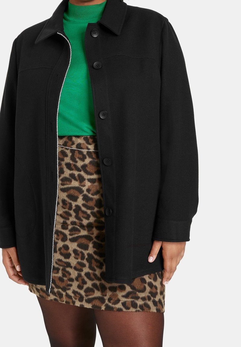 Veste noire boutonnée sur un haut vert vif associée à une mini-jupe à imprimé léopard et des collants foncés portée par une personne sur fond blanc.
