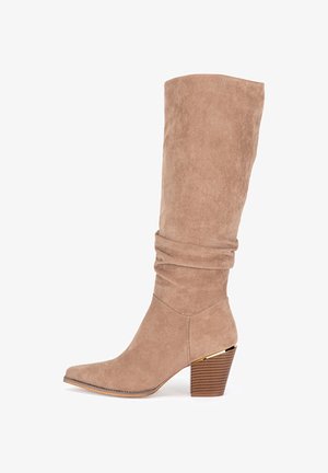 Beżowy zamszowy kozaki za kolano o luźnym fasonie, ze spiczastym noskiem i brązowym, drewnianym, blokowym obcasem z teksturą oraz złotym akcentem.
