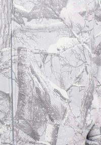 Tela con patrón de camuflaje en tonos grises y rosas, que recuerda a la corteza y ramas de árboles, con una textura suave y detalles visibles de costura.