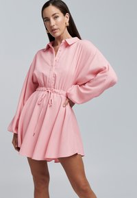 Roze shirtjurk met een kraag, knoopsluiting aan de voorkant, elastische taille en lange, volumineuze mouwen. Glad stof met een zachte textuur.