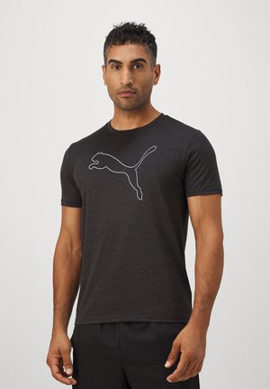 Muškarac nosi pripijenu crnu Puma majicu s bijelim obrubom logotipa Puma, stoji na jednobojnoj svijetlosivoj pozadini.