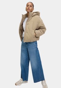 Beige pufferjakke med hætte, der har lynlås foran og trykknapper. Båret med bredbenede blå jeans og lyse sneakers.