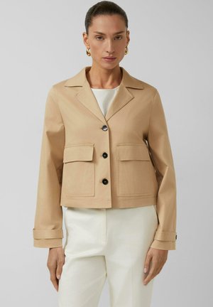 Kobieta nosząca beżową krótką kurtkę z czarnymi guzikami i dużymi przednimi kieszeniami, połączoną z białymi spodniami i złotymi kolczykami kołami.