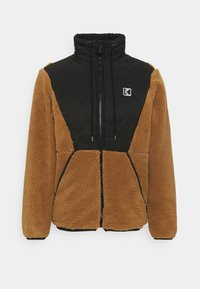 Fleece bunda s hnedou, chlpatou textúrou a čiernym prešívaným panelom na vrchu. Má vysoký golier, zips na uzatváranie a dve zapínateľné vrecká.
