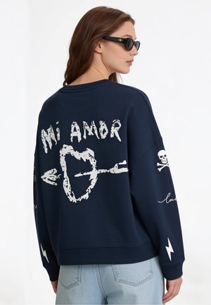 Femme portant des lunettes de soleil, habillée d'un sweatshirt bleu marine orné d'un graphique cœur et flèche "Mi Amor,", tête de mort et os croisés sur la manche, et d'un jean bleu clair.