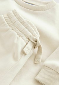 Sudadera de algodón color beige con puños acanalados, que presenta un bolsillo fruncido con cordón. Textura suave y diseño minimalista.