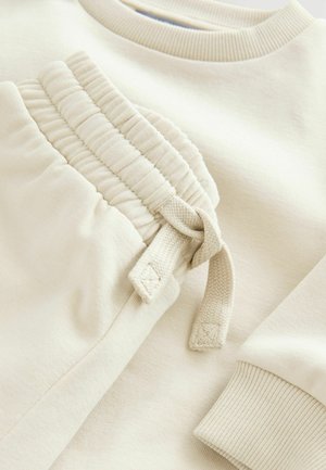 Beige puuvillane dressipluus ribitud mansettidega, mis sisaldab kogutud taskut, millel on lips. Sile tekstuur ja minimalistlik disain.