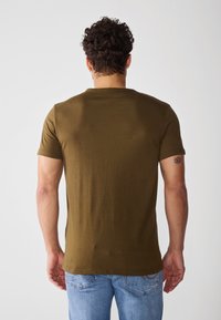 Tricou verde măsliniu cu mâneci scurte, dintr-un material neted, cu un design ajustat și guler rotund, purtat cu blugi de culoare albastru deschis.