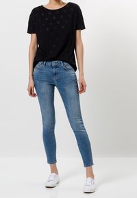 Top de manga corta negro con puntos blancos combinado con jeans ajustados de color azul claro. La modelo lleva zapatillas blancas. Diseño sencillo y ajuste casual.