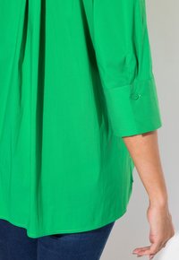 Blusa verde de tejido suave con mangas de tres cuartos, que presenta un detalle de botón en el puño y un corte relajado.