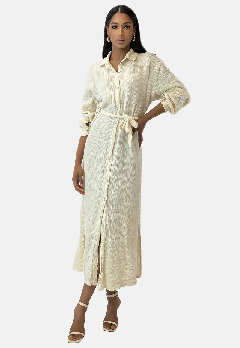Abito camicia lungo color crema, con bottoni, maniche lunghe e tessuto strutturato, cintura in vita e spacco anteriore, abbinato a sandali leggeri con cinturini.