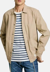 Giacca beige con bottoni, colletto e tasche laterali, indossata sopra una camicia a righe blu e bianche, abbinata a jeans blu.