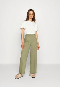 Mulher vestindo uma blusa branca de mangas curtas, calças de cintura alta verdes oliva com cinto, sandálias bege e óculos de sol com lentes âmbar, em pé contra um fundo simples.