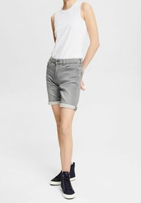 Osoba w białej bezrękawnikowej bluzce, z podwiniętymi szarymi szortami dżinsowymi i czarnymi, wysokimi trampkami, stojąca na białym tle.